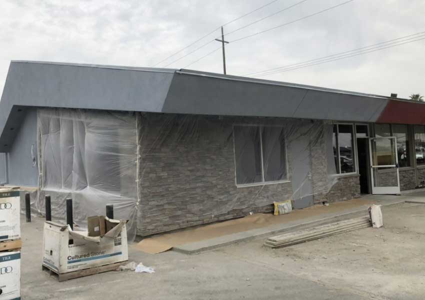 Commercial exterior painting in progress with grey paint being applied to a stone facade building and windows masked off with plastic sheeting, showing materials like 'Cultured Stone' boxes on a pallet